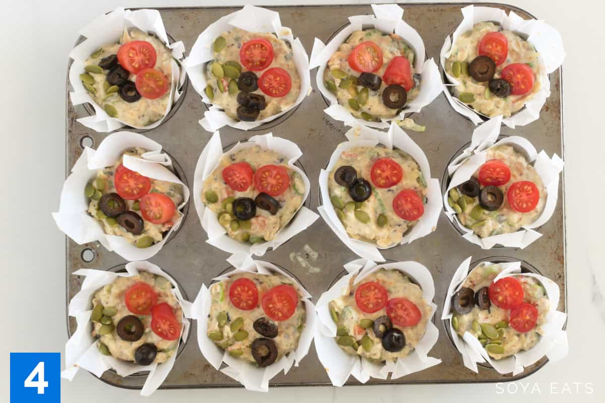 Muffin batter in muffin cases garnished with sliced tomato, black olives and pumpkin seeds.