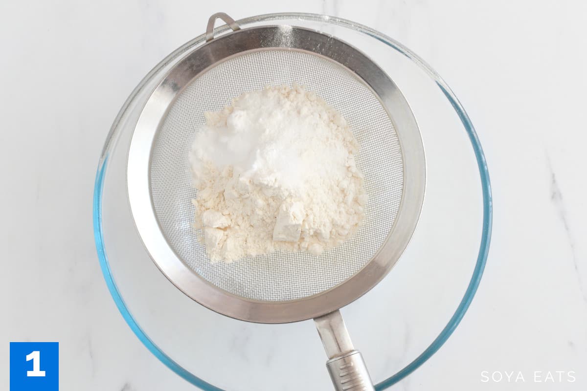 Flour and baking powder in a sieve sitting on top of a mixing bowl.