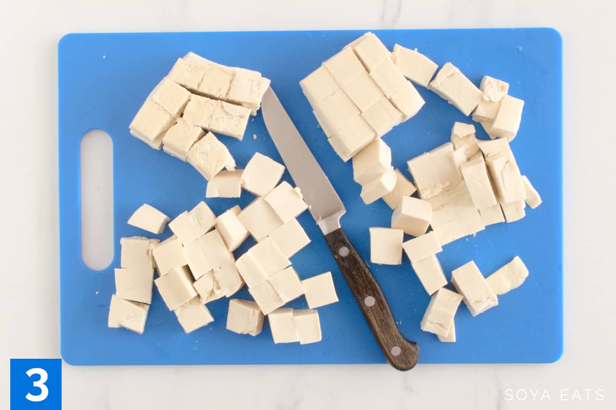 Tofu cut into small cubes on a blue cutting board with a small knife.